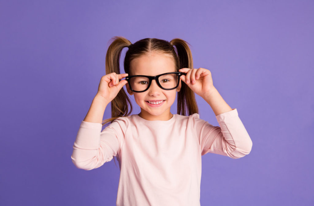 Young girl against a lavender background putting on a pair of glasses.