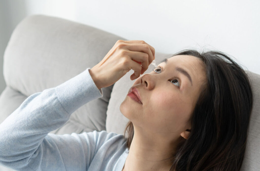 A woman in a sweater leaning backward and looking upwards to apply artificial tears on her right eye.