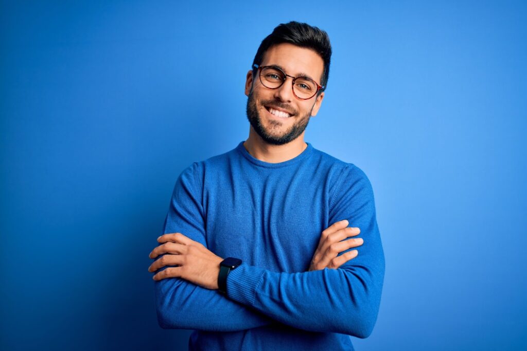 A smiling young man wearing glasses and a blue shirt and standing against a blue background with his arms crossed.