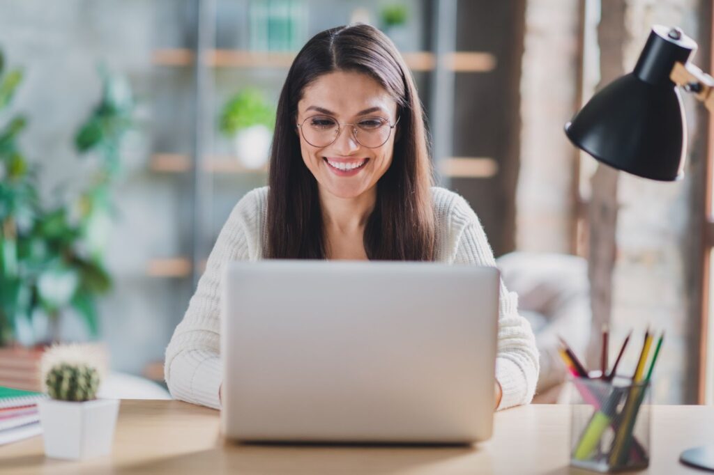 Young woman wearing glasses and smiling while working at a desk on her laptop.