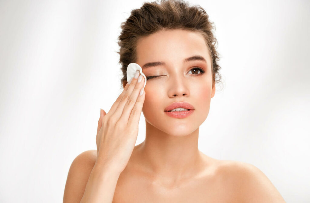 A young woman removing eye makeup on her right eye using a cotton pad with her right hand.