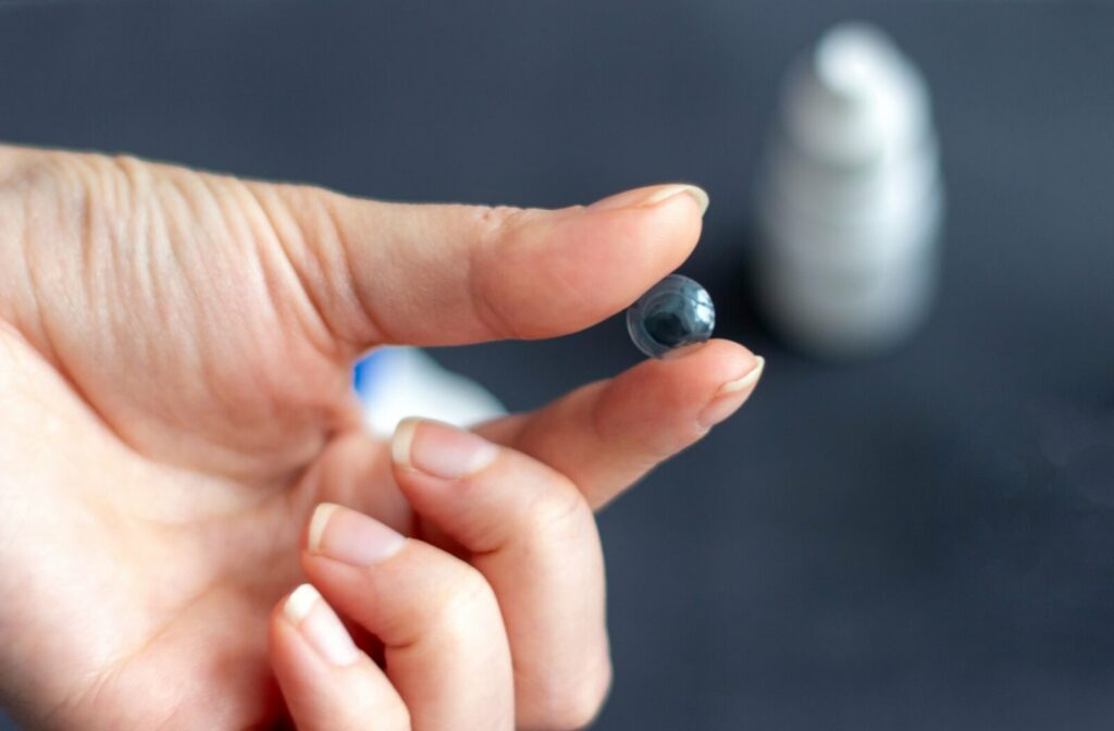 A close up view of a left hand holding an ortho-k contact lens between the thumb and index finger. A bottle of eye drops is out of focus in the background.