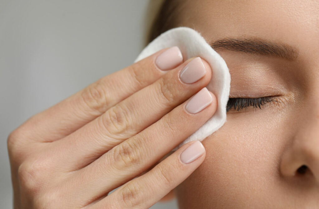 A close-up of one side of a person's upper face. The person is wiping their eye makeup away with a cotton pad.