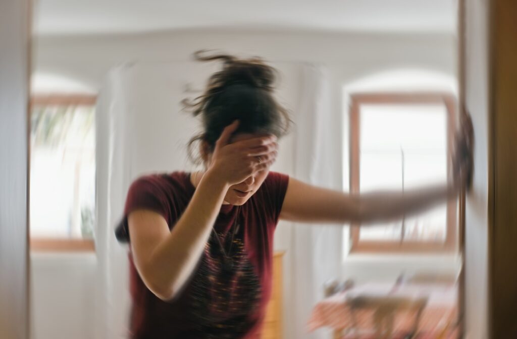 Person with dark hair grips the wall next to them, leaning into it, with their other hand is on their head, shielding their eyes. The environment around them is distorted and blurred.