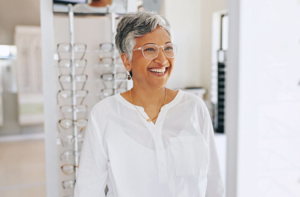 Smiling person with grey hair wearing clear glasses in a bright eyewear store with shelves of frames in the background.