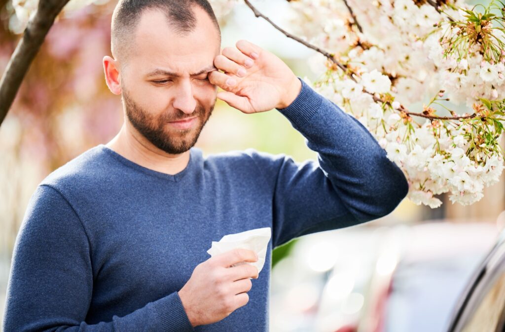 A person standing under a flowering tree rubs their irritated eye.