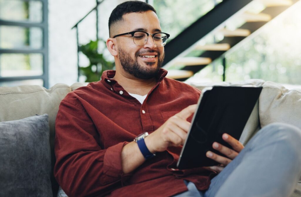 A person wearing glasses sits on their couch at home and scrolls through their tablet.