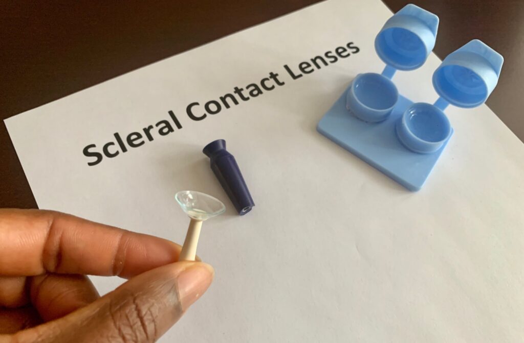 A hand holds a plunger tool that cups a scleral contact lens against a paper that reads "Scleral Contact Lenses.