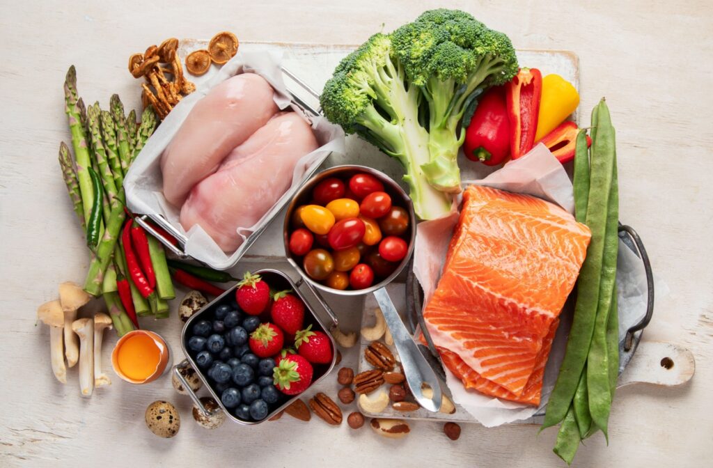 health foods arranged together including chicken breast, salmon, tomatoes, broccoli, berries, and nuts.
