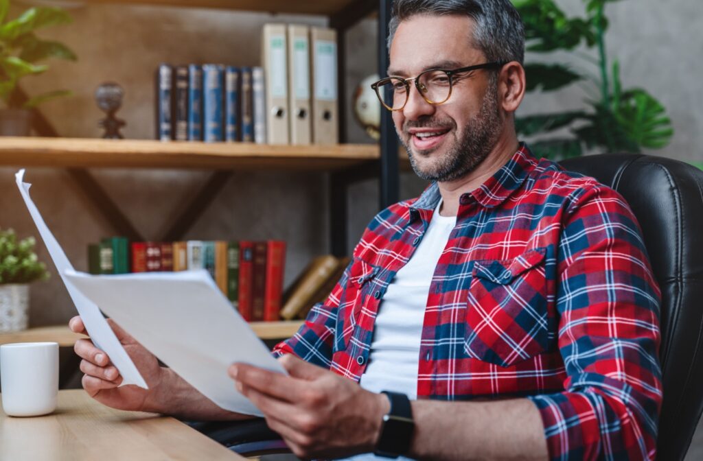 Smiling man in glasses reading documents at his desk with bookshelves in the background.