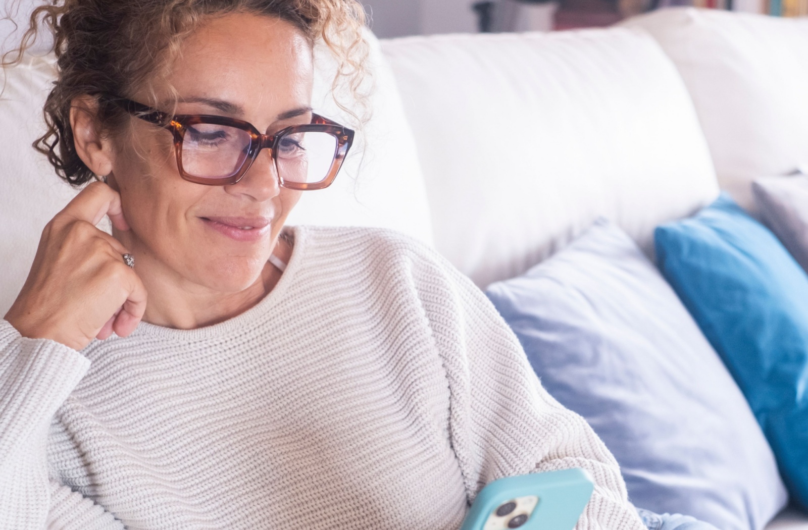 Woman wearing reading glasses sitting on a couch and looking at her smartphone with a relaxed smile.
