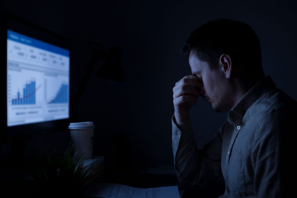 Person in business attire rubbing eyes while working late at computer in dark office with coffee cup nearby, showing symptoms of digital eye fatigue.