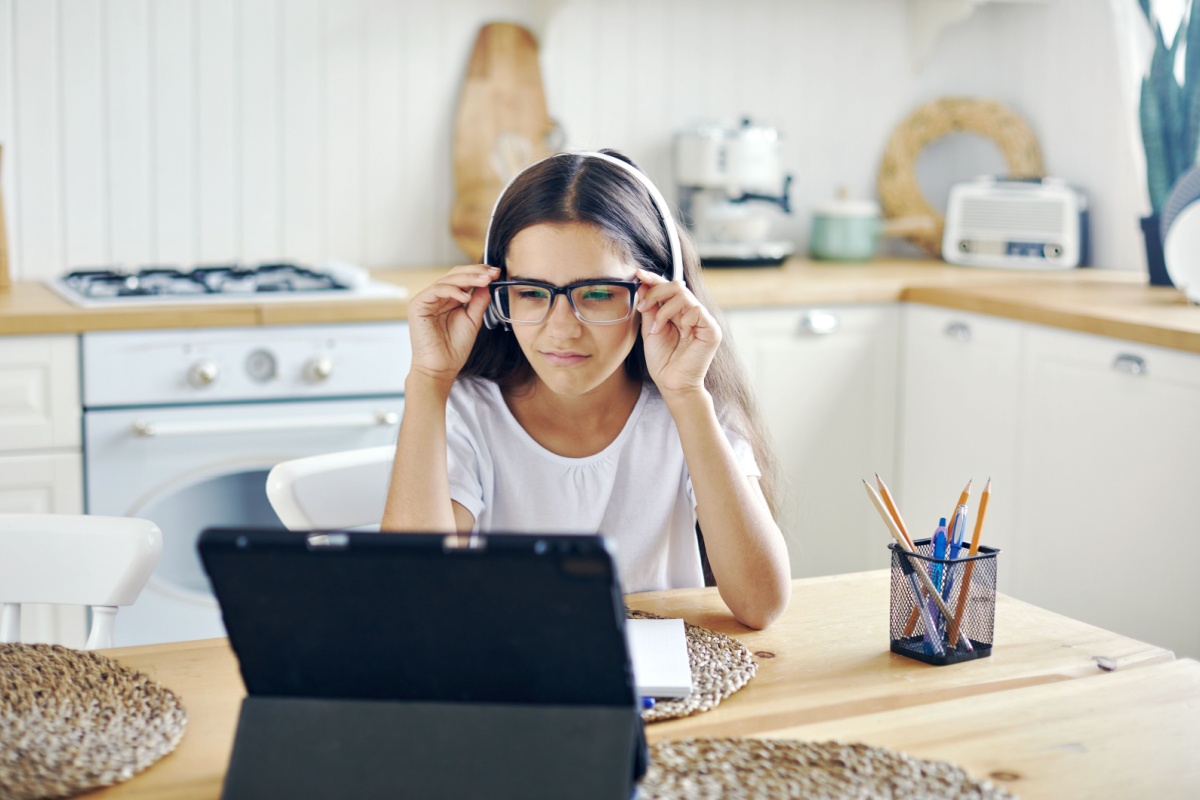 Young girl wearing eyeglasses adjusting frames while doing homework on tablet at kitchen table.